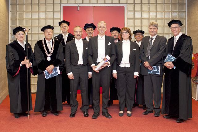 Group photo of eleven people in academic dress and suits, some holding copies of the dissertation, in front of a red wall.